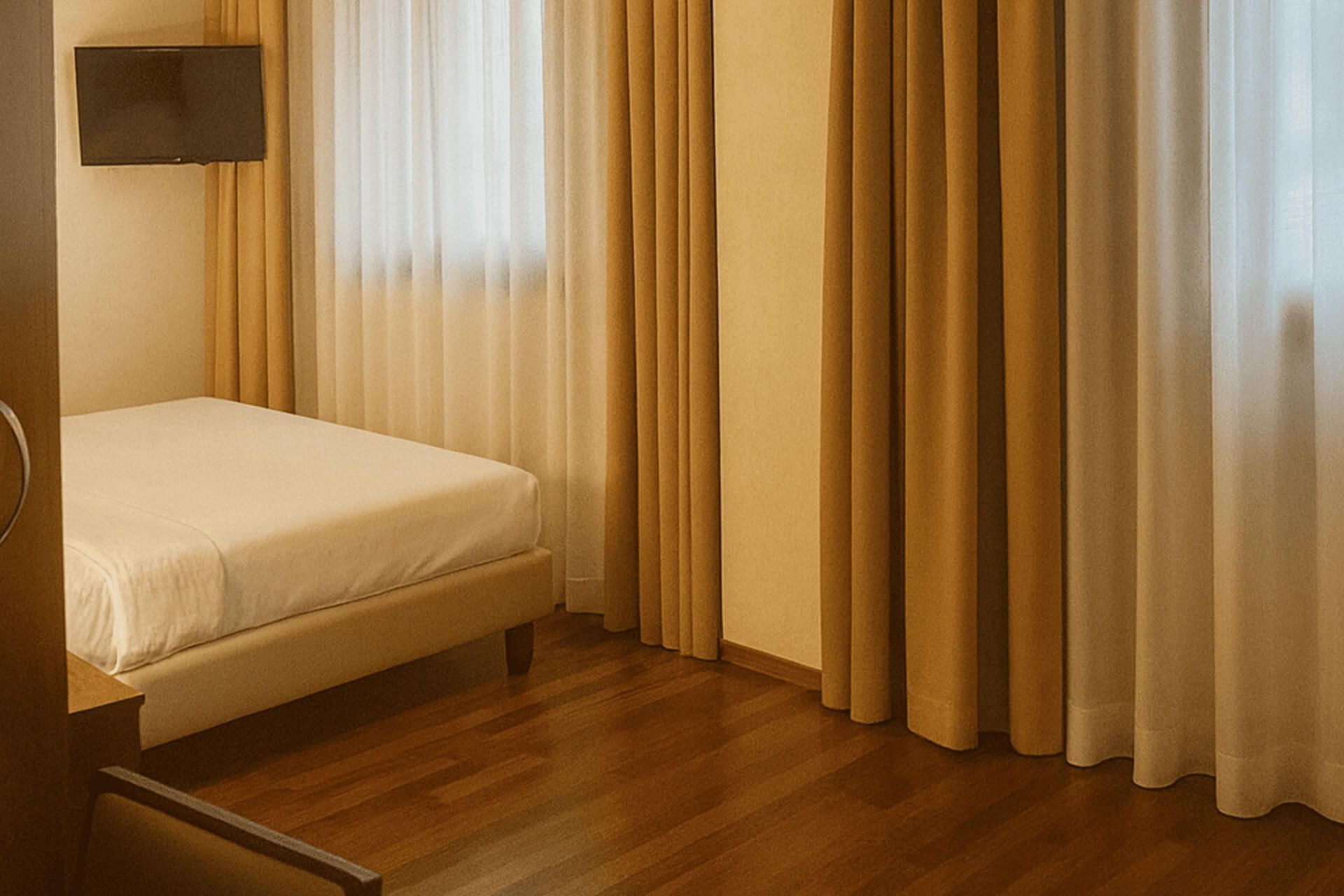 Angolo di camera d’hotel con letto matrimoniale, pavimento in legno, grandi tende color ocra davanti alle finestre e TV a schermo piatto a parete.