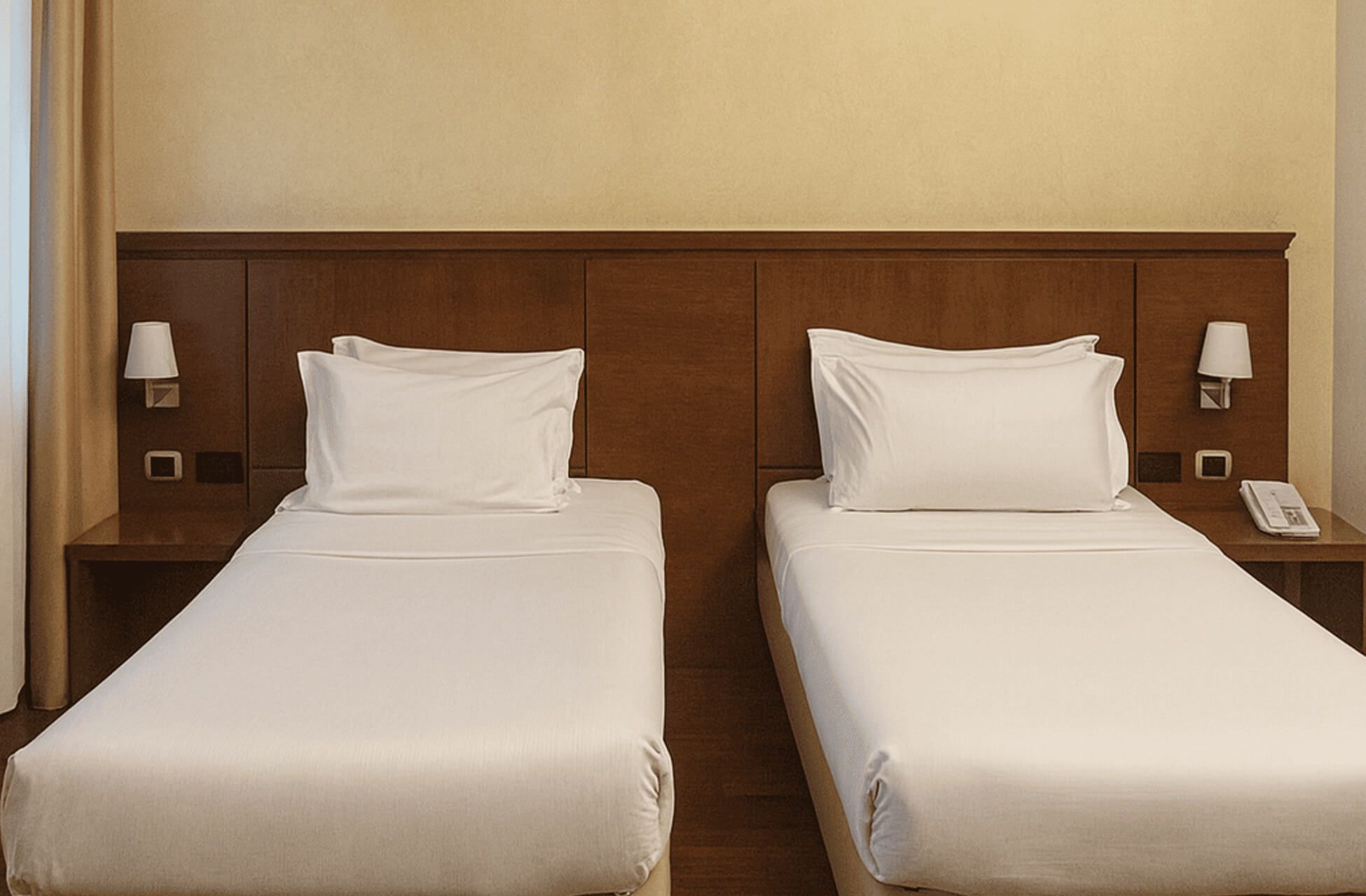 Due letti singoli affiancati di una camera tripla d’hotel, con testata in legno, comodini laterali, lampade da parete e telefono sul lato destro.