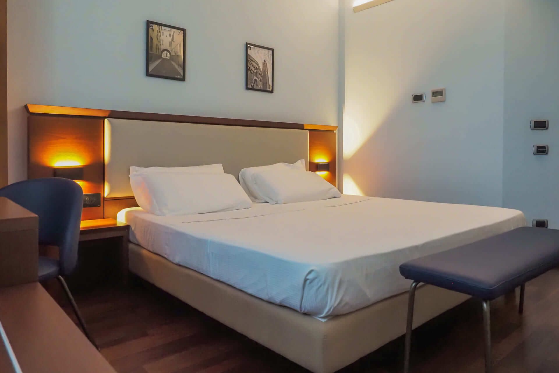 Camera d’albergo moderna con letto matrimoniale, testiera illuminata, panca ai piedi del letto e parete chiara decorata con due quadri.