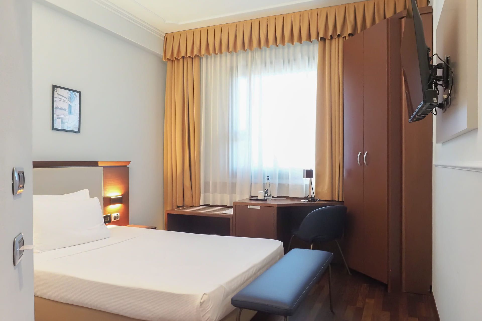 Camera d’albergo compatta con letto alla francese, scrivania sotto una grande finestra con tende dorate, armadio in legno e TV montata a parete.