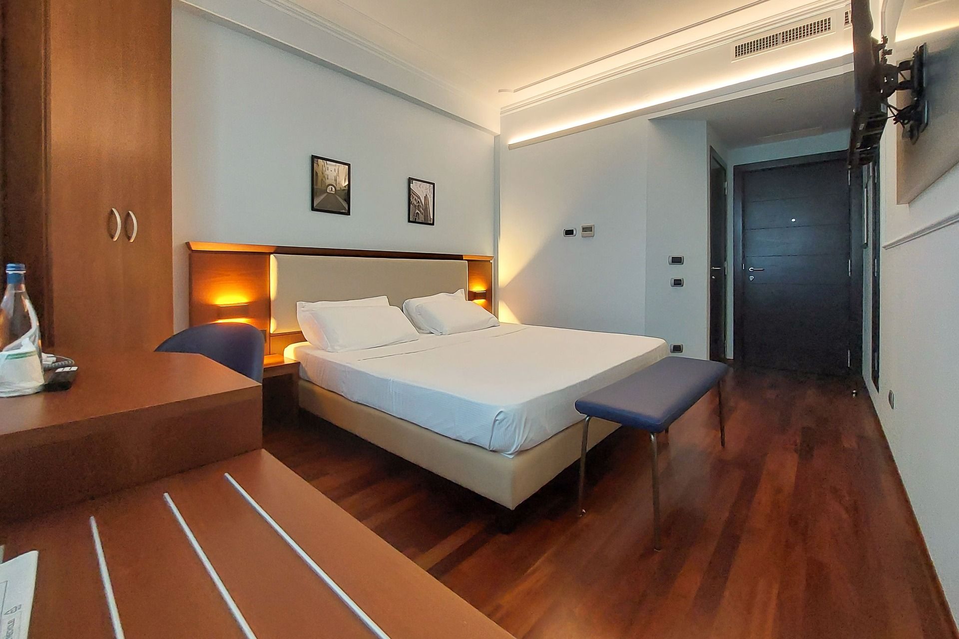 Camera d’albergo moderna con letto matrimoniale, arredi in legno, luci calde dietro la testiera, pavimento in parquet e TV a parete