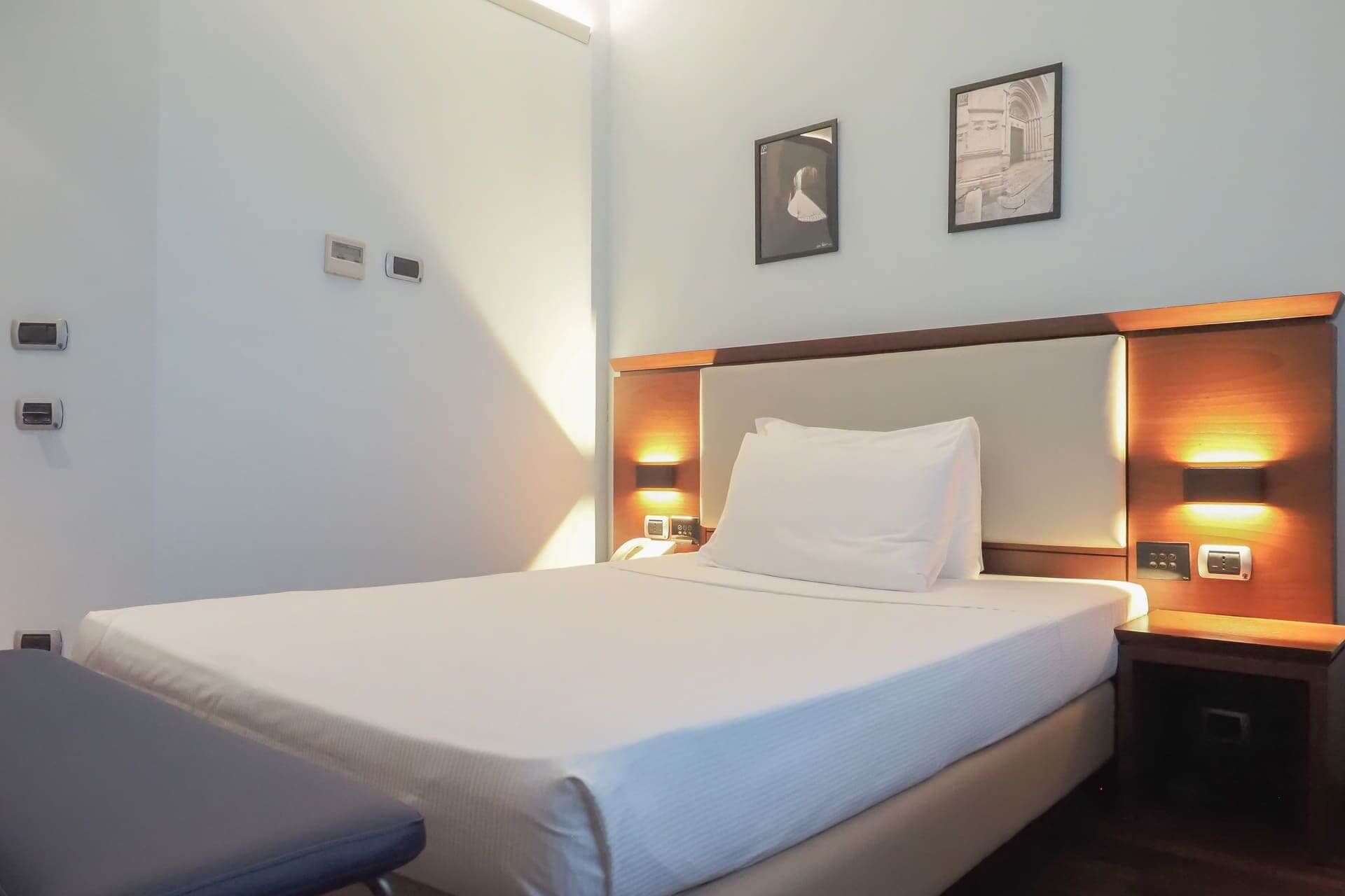 Camera singola con letto alla francese, testiera in legno illuminata, parete chiara decorata con due quadri e finestra con tende color ocra.