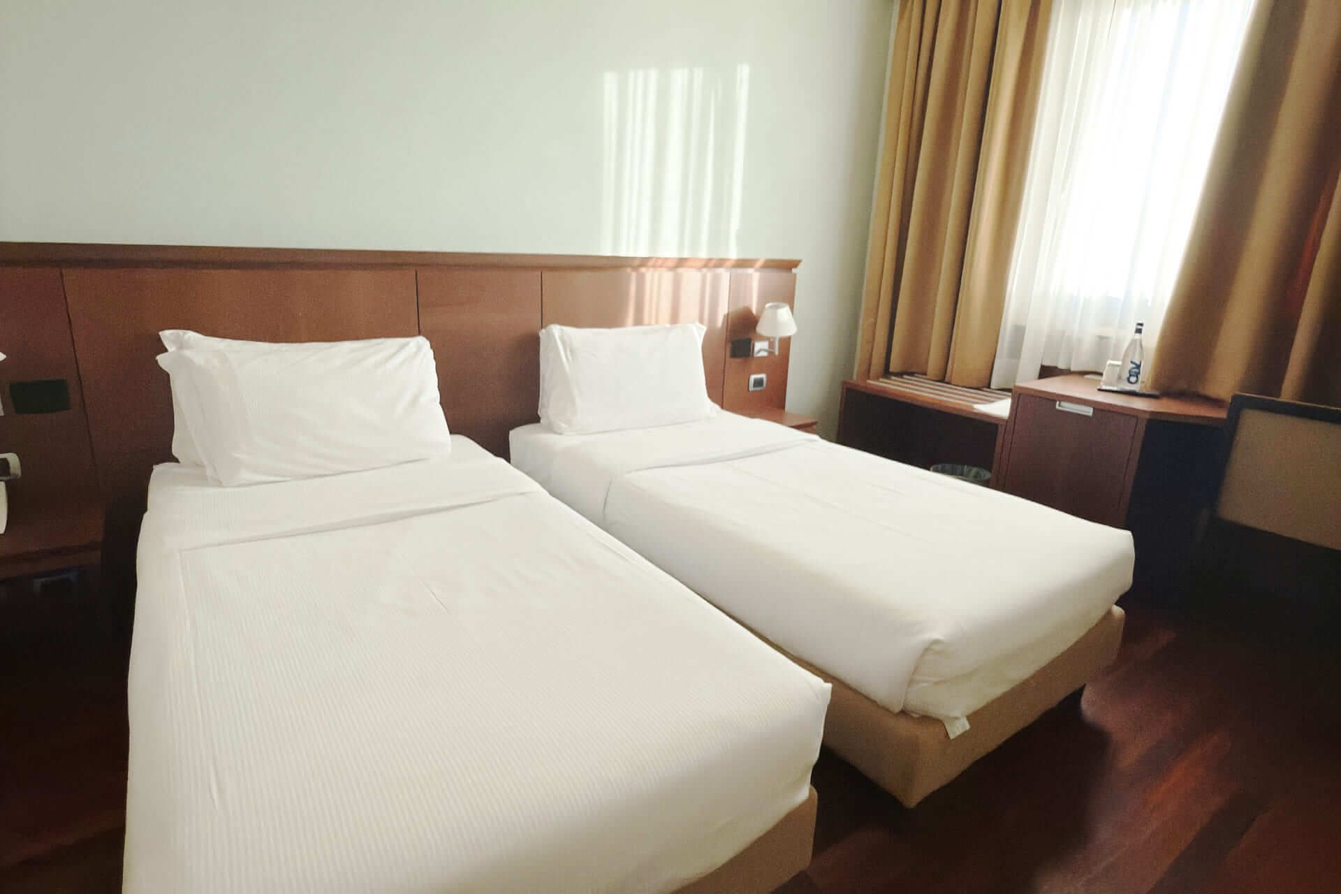 Camera d’albergo con due letti singoli affiancati, pavimento in legno scuro, tende dorate davanti a una finestra luminosa e arredi in legno