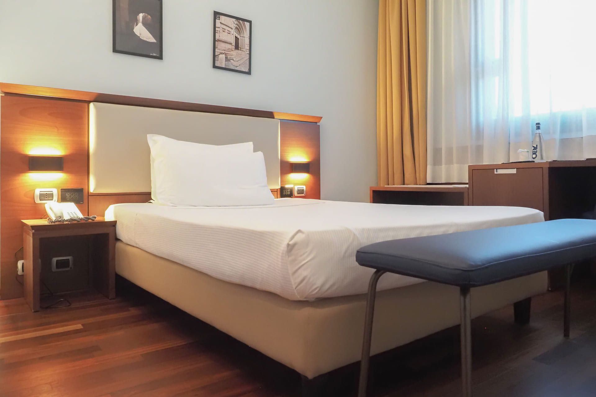 Camera d’albergo luminosa con letto alla francese, luci da parete, pavimento in legno, panca ai piedi del letto e finestra con tende chiare.