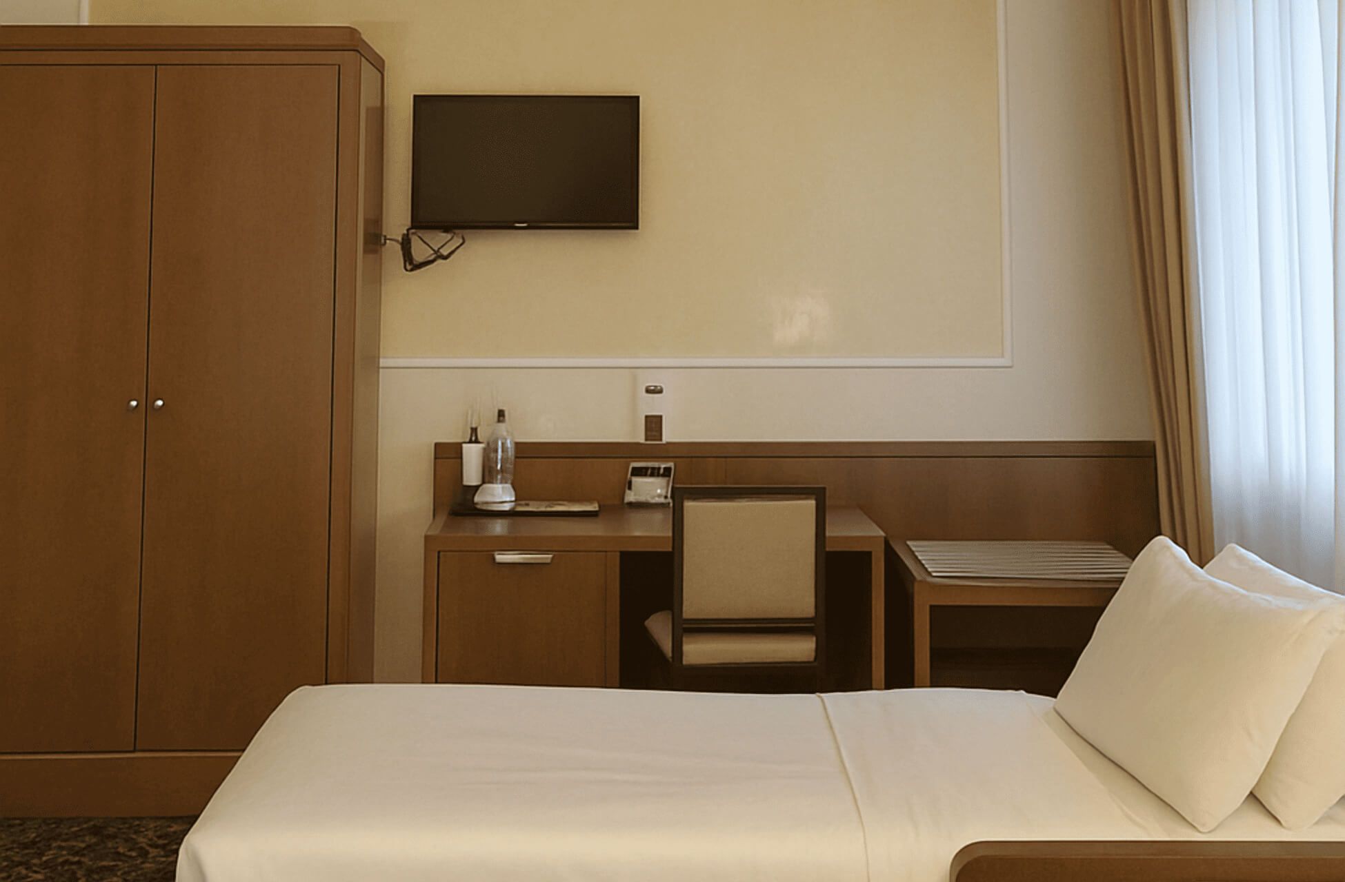 Angolo di camera tripla d’hotel con letto singolo in primo piano, armadio e scrivania in legno con sedia, TV a schermo piatto a parete e finestra con tende chiare.