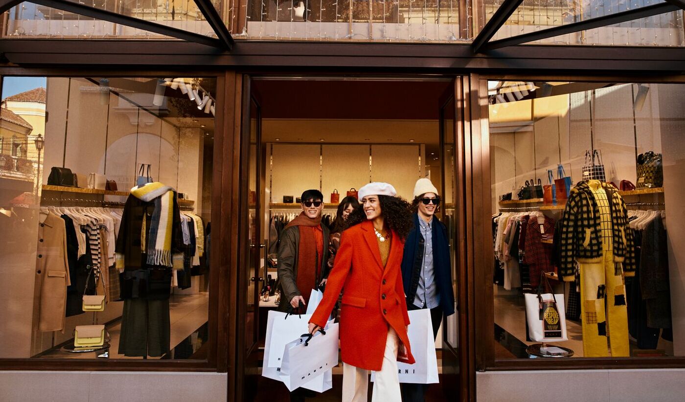 Gruppo di persone eleganti esce da una boutique di moda con grandi vetrine illuminate, indossando cappotti invernali e tenendo borse da shopping, in un contesto urbano raffinato.