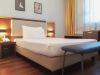 Camera d’albergo luminosa con letto alla francese, luci da parete, pavimento in legno, panca ai piedi del letto e finestra con tende chiare.