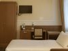 Angolo di camera tripla d’hotel con letto singolo in primo piano, armadio e scrivania in legno con sedia, TV a schermo piatto a parete e finestra con tende chiare.