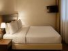 Letto matrimoniale in suite junior d’hotel, con testata in legno, comodino e lampade da parete, TV a schermo piatto e finestra con tende chiare sullo sfondo.