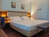 Camera d’albergo moderna con letto matrimoniale, testiera illuminata, panca ai piedi del letto e parete chiara decorata con due quadri.