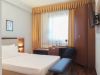 Camera d’albergo compatta con letto alla francese, scrivania sotto una grande finestra con tende dorate, armadio in legno e TV montata a parete.
